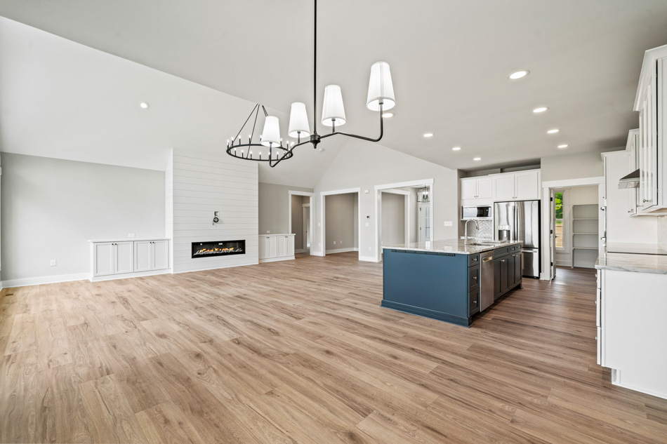 A well-illuminated luxurious house interior design with a fireplace on one side of the wall, a wagon wheel and a linear chandelier, and a wide kitchen island, and a laminate flooring